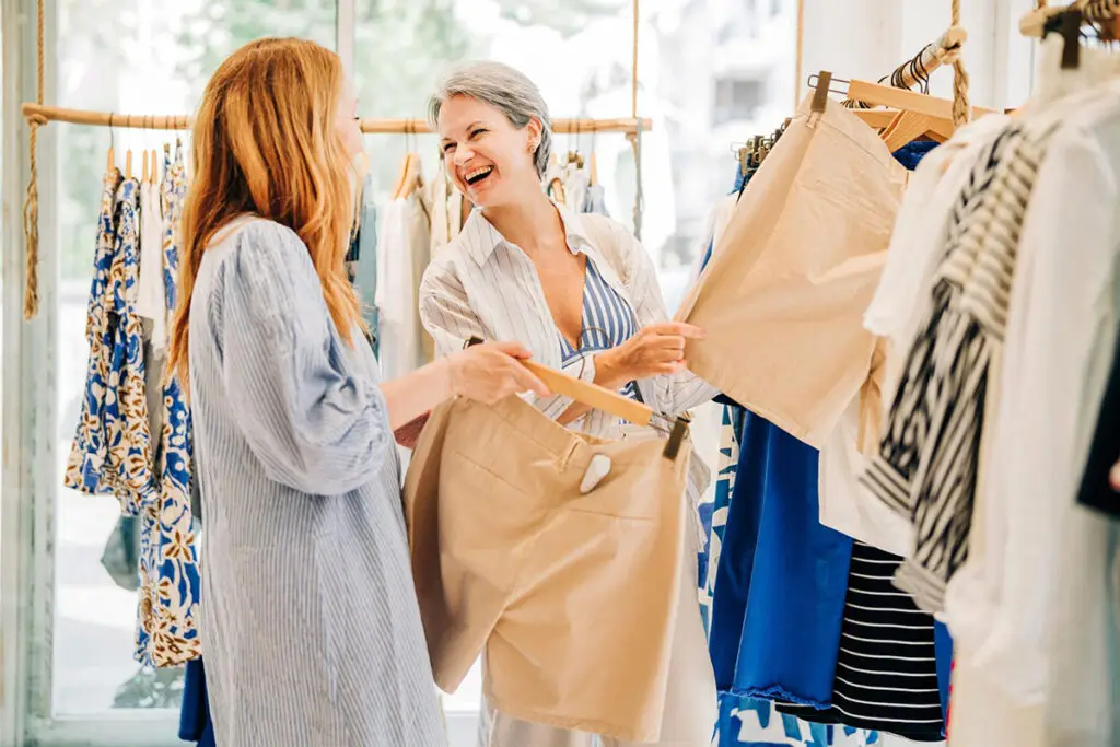 Zwei Frauen lachen in der Boutique zwischen Kleiderstangen, eine hält eine beige Shorts hoch